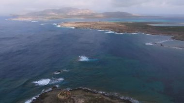 Sardunya sahilinin turkuvaz suları, kumlu kıyı şeridi ve plaja gidenlerle hava manzarası. Kıyı şeridinde bir yol, park edilmiş arabalar ve yemyeşil alanlar yer alıyor..