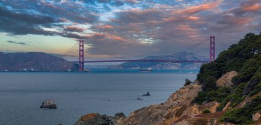 İkonik Golden Gate Köprüsü San Francisco Körfezi boyunca uzanır, kırmızı turuncu renkler mavi sulara ve yemyeşil alana karşıdır. Marin County tepeleri görülebilir..