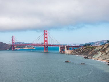 Golden Gate Köprüsü, Uluslararası Turuncu rengiyle, arka planda parçalı bulutlu gökyüzü ve yuvarlanan tepeler ile San Francisco Körfezi boyunca uzanır..