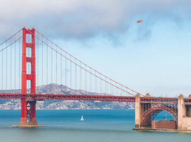 Golden Gate Köprüsü San Francisco Körfezi boyunca uzanır. Altında bir yelkenli ve üstünde bir helikopter vardır. Marin County tepeleri arka planda sisli.