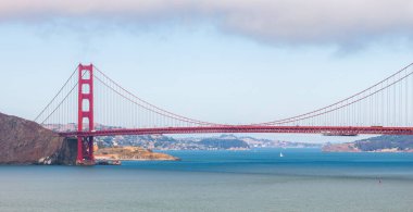 Golden Gate Köprüsü San Francisco Körfezi boyunca uzanır, turuncu rengi mavi sular ve uzak tepelerle çelişir. Yelkenliler ölçek ve aktivite ekler.