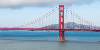 Golden Gate Köprüsü kırmızı turuncu rengiyle San Francisco Körfezi boyunca uzanır. Marin County tepeleri ve yelkenlileri bulutlu bir gökyüzünün altında görülebilir..