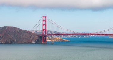 Golden Gate Köprüsü San Francisco Körfezi boyunca uzanır, solda Marin Burunları ve bulutların altında şehir manzarası bulunur..