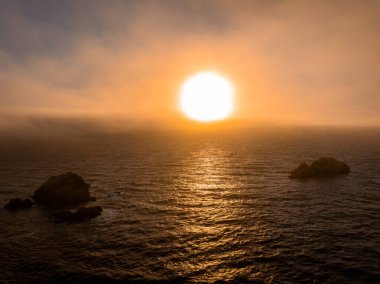 Görüntü Pasifik Okyanusu 'nun üzerinde gün batımını gösteriyor, suyun üzerinde altın bir ışık var. Ön planda, sisli bir gökyüzünün altında iki kaya oluşumu var..