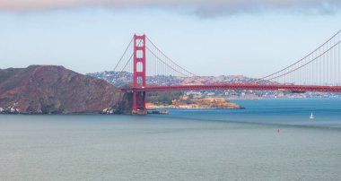 Golden Gate Köprüsü, uluslararası turuncu rengiyle San Francisco Körfezi boyunca uzanır. Mavi sularda yüzen bir yelkenli, Marin County tepelerinde görünür..