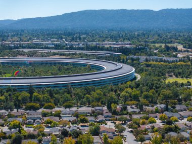 Görüntü, Cupertino, California 'daki Apple Parks' ın yeşil, yerleşim alanları ve yuvarlak tepelerle çevrili dairesel mimarisinin havadan görüntüsünü yakalıyor..