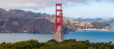 Golden Gate Köprüleri, yuvarlanan tepelere ve parçalı bulutlu gökyüzüne karşı yükselen kırmızı turuncu kuleler, San Francisco Körfezi 'nden görünen teknelerle.
