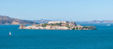 Tarihi cezaevi ve gözlem kulesiyle Alcatraz Adası San Francisco Körfezi mavi sularıyla çevrilidir. Bir yelkenli ve Marin Burunları görünüyor..