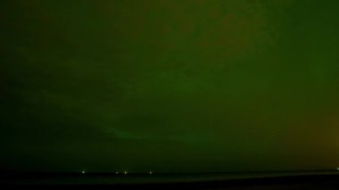 Parlak yeşil aurora borealis, durgun su üzerinde gece gökyüzünü aydınlatır. Parlak bir meteor ya da uydu olay yerinde hızla ilerler..