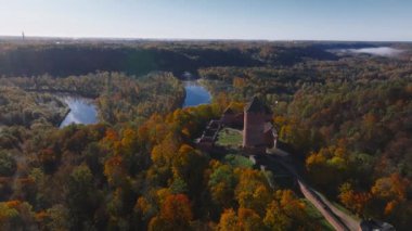 Sigulda, Letonya 'nın hava manzarası, açık mavi gökyüzü altında canlı sonbahar yaprakları ve Turaida Kalesi, mevsimsel güzellikleri vurguluyor..