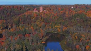 Sigulda, Letonya 'nın hava manzarası, açık mavi gökyüzü altında canlı sonbahar yaprakları ve Turaida Kalesi, mevsimsel güzellikleri vurguluyor..