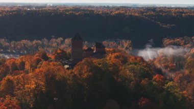 Sigulda, Letonya sonbahar manzarasında canlı kırmızı, turuncu ve sarı yapraklar bulunur. Sis ormanın üzerinde süzülür, dingin ve mistik bir atmosfer yaratır..