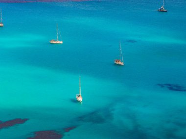 Turkuaz sular, İtalya 'nın Sardinya kıyılarında demirlemiş yelkenlileri çevrelemektedir. Mavi renklerin gradyanı denizin enginliğini ve sükunetini vurgular..