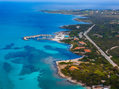 Sardunya, İtalya 'nın havadan görünüşü, turkuaz bir deniz, tekneli küçük bir marina, yemyeşil ve dolambaçlı bir yol boyunca dağınık binalar..