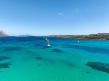 Turkuaz Akdeniz suları, demirli yelkenliler, yemyeşil, yumuşak tepeler ve parlak mavi bir gökyüzünün yer aldığı Sardinya kıyılarının havadan görünüşü..