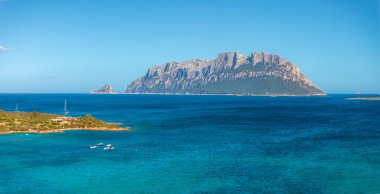 Tavolara Adası 'nda dramatik kireçtaşı uçurumlar ve turkuaz sular bulunur. Küçük tekneler açık mavi gökyüzünün altında yemyeşil kıyı şeridinde demirli..