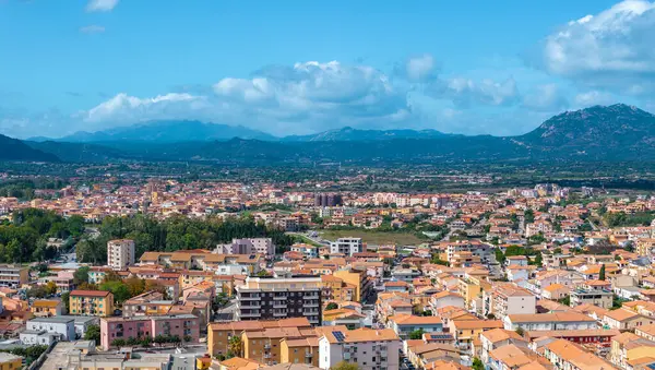 Olbia, Sardinya 'nın havadan görünüşü, terasotta çatısı olan binalar, yemyeşil yemyeşil ve parlak mavi gökyüzü altındaki dağlar, kentsel ve doğal elementleri gözler önüne seriyor..