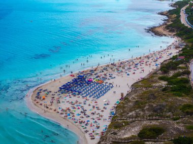 Renkli şemsiyeler, güneşlenenler, turkuaz Akdeniz suları ve dolambaçlı bir kıyı yolunun yer aldığı İtalya 'daki hareketli bir Sardunya plajının havadan görünüşü.