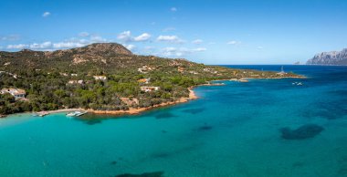 İtalya 'nın Sardinya kentindeki turkuaz bir körfezin havadan görünüşü. Etrafı yeşillik ve konut binalarıyla çevrili. Tekneler sakin suları arka planda dağlarla dolduruyor..