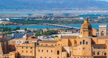 Terracotta binalı Cagliari manzarası, önemli bir kubbe ve sanayi bölgesine giden bir köprü. Dağlar ve su manzaralı bir zemin oluşturuyor..