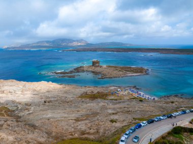 Sardinya 'daki küçük bir adada Torre della Pelosa' nın hava manzarası, turkuaz sular, kumlu plaj ve parçalı bulutlu bir gökyüzü altında engebeli manzarayla çevrili..