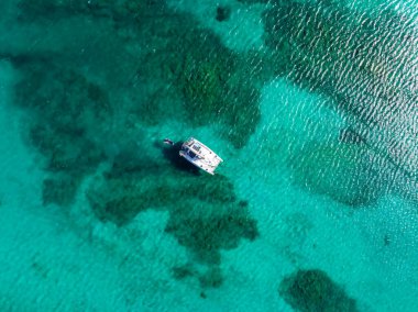 A boat floats on clear turquoise waters near Sardinia, Italy, surrounded by patches of darker blue indicating deeper areas or underwater vegetation.