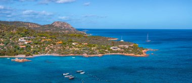 Canlı mavi deniz, küçük tekneler ve yatların yer aldığı Sardinya kıyı şeridinin havadan görünüşü. Yeşillik arka planda engebeli bir tepe ile kıyı şeridi çiziyor..