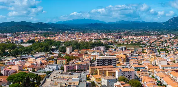 Olbia, Sardinya 'nın hava perspektifi, terakotta çatılı binalar, yemyeşil, yuvarlanan tepeler ve bulutlu bir gökyüzünün altındaki uzak dağlar.