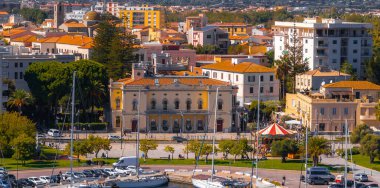 Görüntü, Olbia, Sardinya 'da, klasik mimarisi olan tarihi bir bina, rıhtımlı tekneleri olan bir marina ve renkli konut binalarının yer aldığı canlı bir kent görüntüsü yakalıyor..