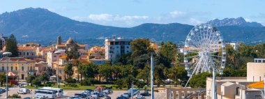 Olbia, Sardinya 'da büyük bir dönme dolap, renkli binalar ve kubbe bir yapı bulunur. Yeşillik ve engebeli dağlar manzaralı bir zemin oluşturur..