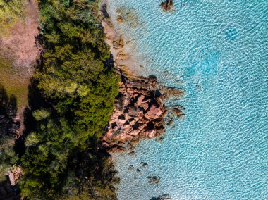 Sardunya kıyılarının yemyeşil, turkuaz berrak suları ve denize doğru uzanan kızıl kahverengi kayaları gösteren hava manzarası..
