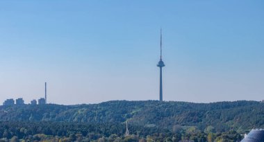 Vilnius TV Kulesi, yemyeşil ormanlarla çevrili açık mavi bir gökyüzüne karşı dimdik ayakta duruyor. Yüksek binalar ve bir baca uzaktan görülebilir..