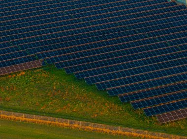 Bir çitle çevrili, hafif eğimli çimenlik bir alanda sıralanmış güneş panelleri. Yeşil çimenler, yenilenebilir enerji kullanımını gösteren koyu mavi panellerle çelişiyor..