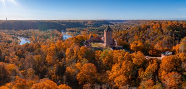 Aerial view of Turaida Castle in Sigulda, Latvia, surrounded by vibrant autumn foliage. The Gauja River winds through the landscape under a clear blue sky. clipart