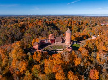 Turaida Castle in Sigulda, Latvia, is seen from above, surrounded by a forest in autumn hues. The red brick castle contrasts with the colorful leaves and blue sky. clipart