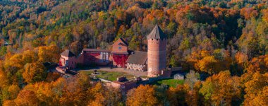 Turaida Castle in Sigulda, Latvia, is seen from above, surrounded by vibrant autumn trees. The red brick walls and conical tower contrast with the colorful forest. clipart