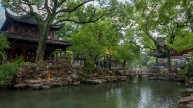 Ziyaretçiler, Yu Garden 'daki taş patikalarında yürürler, etrafı karmaşık ahşap oymalar, kıvrımlı çatılar ve canlı yeşillikleri yansıtan sakin bir gölet ile çevrilidir..