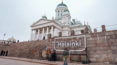 Helsinki Katedrali 'nin neoklasik mimarisi ve yeşil kubbeleri, insanlar basamaklarda dinamik bir şekilde hareket ederken, yukarıda bulutlu bir gökyüzü varken göze çarpıyor..