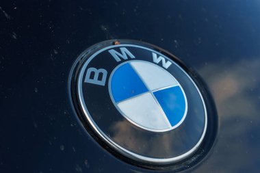 Görüntü, koyu renk bir araç yüzeyindeki BMW logosunu gösteriyor. Logoda gümüş harflerle çevrili mavi ve beyaz daireler yer alıyor. Toz zerreleri görünür..