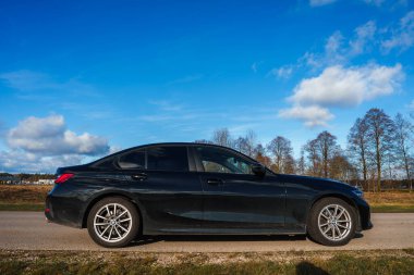 Parlak, siyah bir BMW 320 kırsal bir yolda açık mavi gökyüzünün altında dağınık bulutlarla parkedilmiştir. Çıplak ağaçlar yol boyunca uzanır, bu da sonbaharın sonları ya da ilkbahar başları anlamına gelir..
