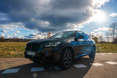 Bir BMW X4 asfalt bir alana park edilmiştir, güneş ışığıyla işaretlenmiştir. Arkaplanda açık bir alan, seyrek ağaçlar ve parçalı bulutlu bir gökyüzü var..