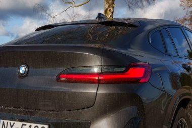 Görüntü bir BMW X4 'ün arka bölümünü yakalıyor, arka lambasını ve logosunu vurguluyor. Bir ağaç ve parçalı bulutlu bir gökyüzü görülebilir..