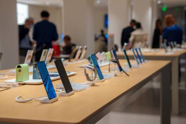 İyi aydınlatılmış, minimalist bir elektronik mağazasındaki parlak ahşap bir masada çeşitli akıllı telefonlar sergileniyor. Arka planda insanlar görünür..