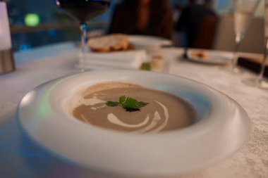 Beyaz bir kasede maydanozla süslenmiş kremalı bir çorba, bir bardak kırmızı şarap ve ekmek ile masaya konmuş, rafine bir yemek atmosferi olduğunu gösteriyor..