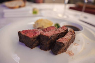 A plated dish features three slices of medium rare steak with a seared exterior, accompanied by mashed potatoes, green garnish, and a side of sauce. clipart