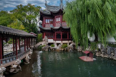Görüntüde Şangay 'daki Yuyuan Bahçesi yer alıyor, ters dönmüş saçaklı bir pavyon, göl üzerinde taş bir köprü ve sulu bir söğüt ağacı da dahil olmak üzere yemyeşil bir köprü yer alıyor..
