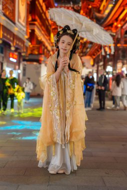 Geleneksel Çin giysileri içindeki bir kişi Şangay 'ın eski kasabasında, süslü ahşap yapılar ve parlak ışıklarla çevrili canlı bir caddede bir güneş şemsiyesi tutuyor..