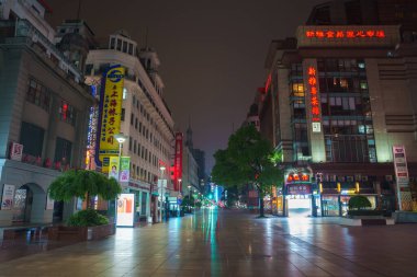 A bustling Shanghai street at night features modern and traditional buildings with neon signs. Wet pavement reflects colorful lights, and trees line the street. clipart