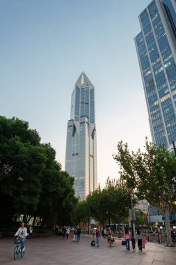 Görüntü, Şangay 'daki Jin Mao Kulesi' ni gösteriyor. Yeşil bir alanda yürüyen ve bisiklet süren insanlar, açık bir gökyüzünün altında, yumuşak ışıklandırmayla..