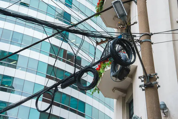 Karmaşık elektrik kabloları, modern cam bir binaya dayanan sokak lambasıyla bir direğin etrafında dönüyor. Balkonlardaki kırmızı çiçekler şehir sahnesine renk katar.